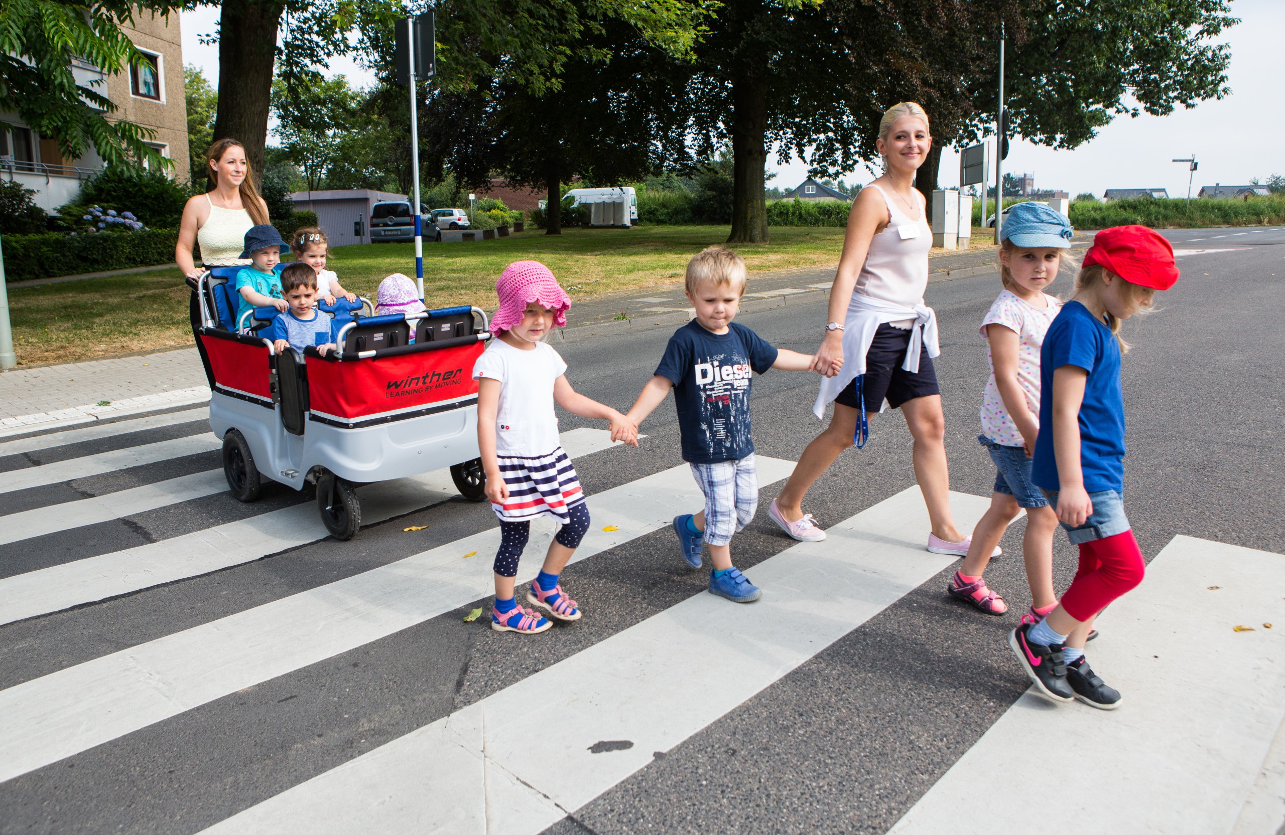 Mehrere Kinder und zwei Erwachsene überqueren zum Teil in einem Rollwagen einen Zebrastreifen