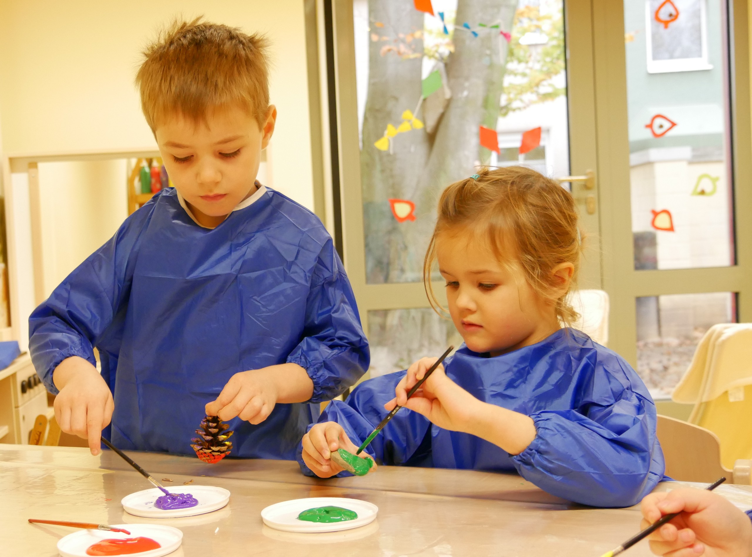 Zwei Kinder bemalen konzentriert Tannenzapfen mit bunter Farbe