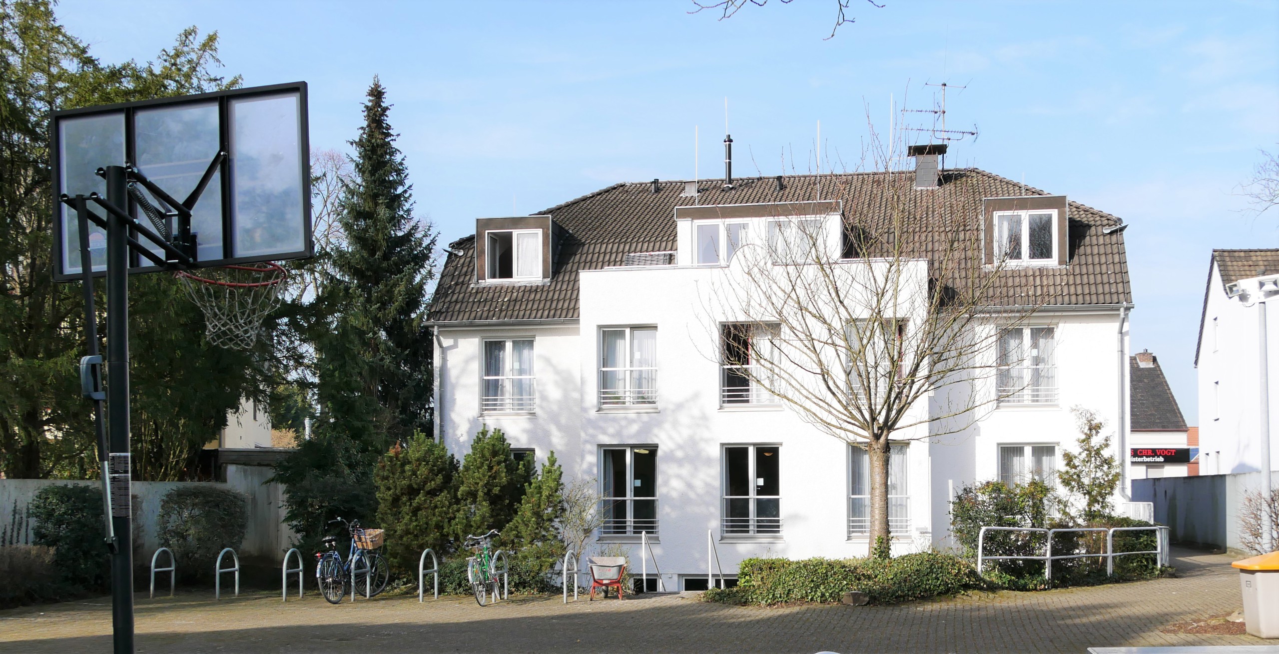 Zweistöckiges weißes Gebäude mit ausgebautem Dachgeschoss, Außenanlage und Basketballkorb